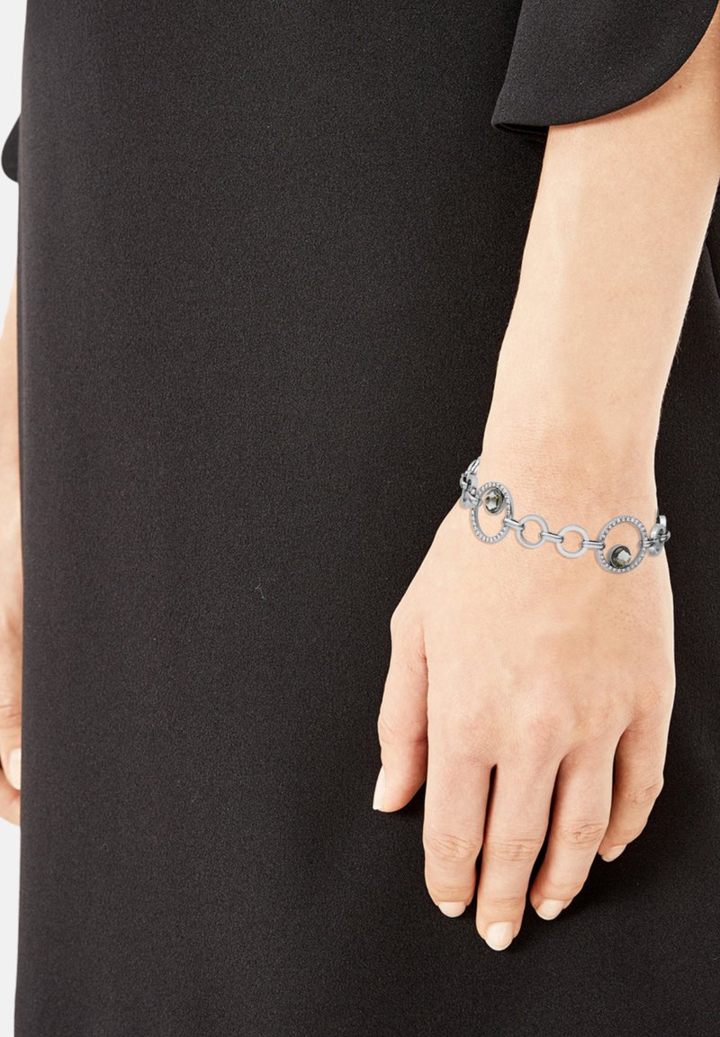 Pulsera de cadena de plata con eslabones circulares y dos piedras oscuras, llevada en una mano sobre un fondo negro sólido.