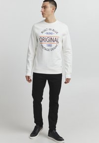 Felpa bianca con stampa circolare che presenta "ORIGINALE" e "1993" in rosso scuro e blu. Abbinata a jeans neri slim-fit e sneaker nere.