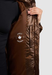 Brūna šķūta jaka no spīdīga materiāla. Tajā ir logotipa uzlīme un sānu kabata. Gluda tekstūra ar elegantu dizainu.