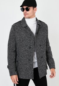 Cappotto a motivo houndstooth bianco e nero con colletto strutturato, bottoni doppi e tasche laterali, indossato sopra un dolcevita bianco.