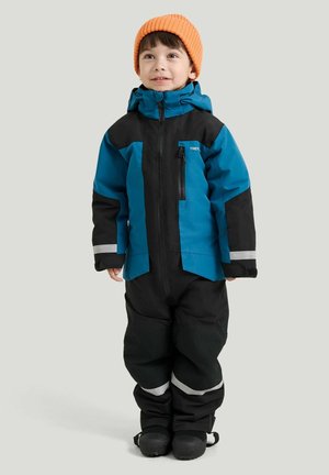 Barn står iført en blå og sort vinterdragt med reflekser og en orange strikhue, kigger let opad og smiler.