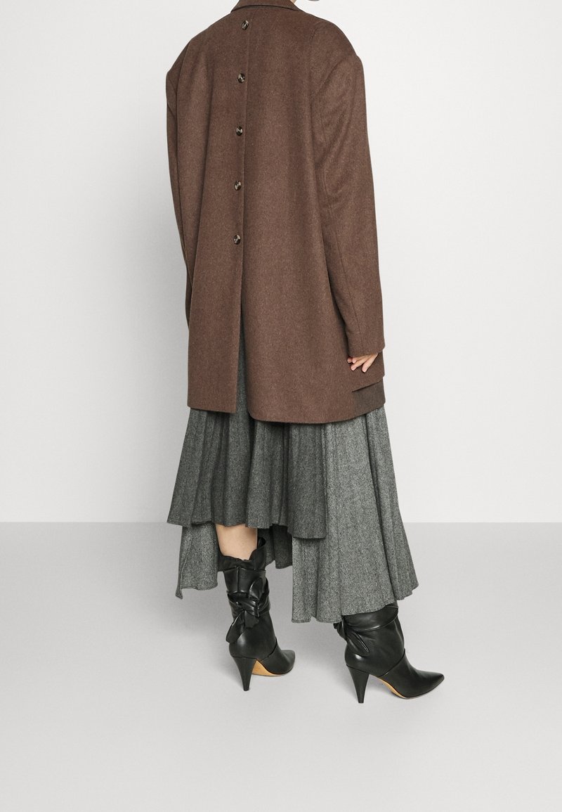 Personne portant un manteau marron boutonné, une jupe midi plissée grise et des bottines noires à talons, debout devant un fond blanc uni.
