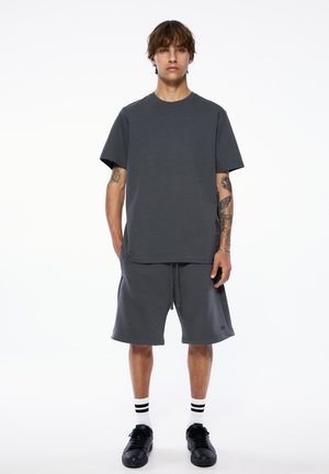 Junger Mann trägt ein dunkelgraues, übergroßes T-Shirt und passende Shorts, mit Tätowierungen an den Armen, weißen Socken mit schwarzen Streifen und schwarzen Turnschuhen.