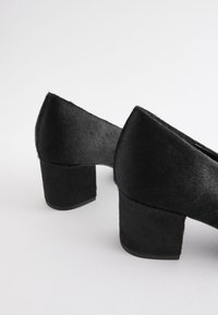 Par svart, midjeklackade skor med en texturerad, pälsliknande yta mot en vit bakgrund, visande bak- och sidovy.