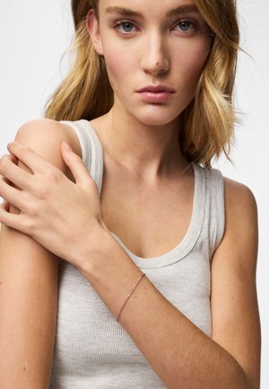 Lichtgrijze geribbelde tanktop, model toont een zilveren kettingarmband om de blote pols, met zachte golven blond haar die het gezicht omlijsten.
