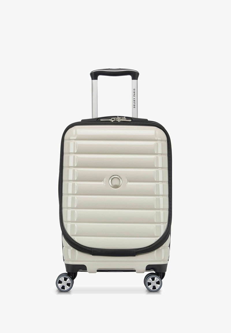 Valise rigide crème avec un design rainuré, accents noirs, une poignée télescopique et quatre roulettes pivotantes pour la mobilité.