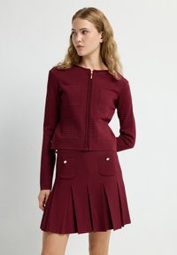 Chaqueta de punto burdeos con cremallera frontal y diseño texturizado, combinada con una falda plisada con bolsillos con botones dorados.