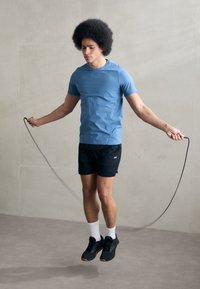 Mann som hopper tau, iført en tettsittende blå t-skjorte, svarte shorts, hvite sokker og svarte sko med brun såle, mot en betongbakgrunn.
