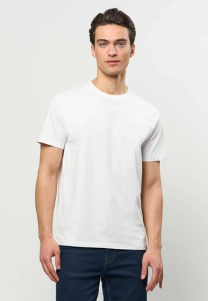 Noormees tume juustega, kandes lihtsat valget lühikese varrukaga t-särki ja tumedat sinist teksapükse, hele taust.