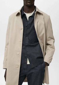 Trench coat in cotone beige con colletto e cinque bottoni frontali, indossato sopra una camicia in denim scuro. Dettagli delle cuciture visibili e una tasca.