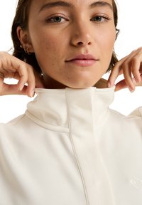 Haut blanc à col montant en tissu doux, avec des détails de couture subtils et un petit logo sur la poitrine. Des mains ajustent le col.