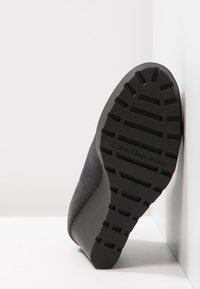 Suola in gomma nera di una scarpa con profondi battistrada, abbinata a una tomaia in suede grigio. Il marchio "Calvin Klein Jeans" è visibile sulla suola.