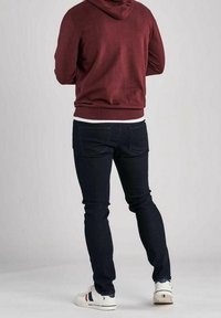 Sweat à capuche bordeaux avec une poche kangourou à l'avant, associé à un jean denim foncé et des baskets blanches avec des rayures colorées. Vue aérienne par l'arrière.