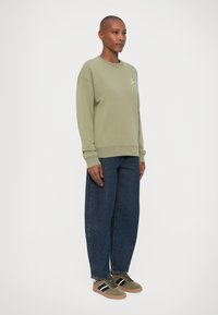 Sudadera verde oliva con cuello redondo, combinada con pantalones de denim azul sueltos y zapatillas verdes con rayas negras y textura de ante.