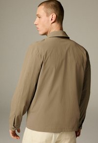 Camisa beige de manga larga hecha de una tela liviana, con cuello, detalles en los puños con botones y un corte relajado. Vista trasera mostrada.
