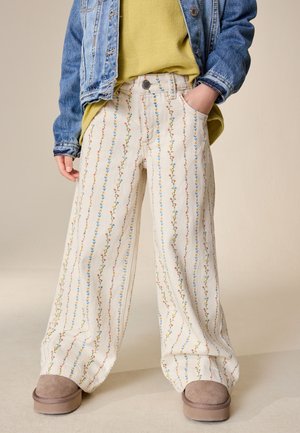 Bambino che indossa pantaloni crema a gamba larga con strisce floreali verticali, un maglione giallo, una giacca di denim blu e scarpe marroni senza lacci.