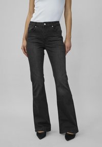 Persoană îmbrăcată cu blugi negri cu talie înaltă și pantaloni evazați, purtând pantofi negri cu vârf ascuțit, asortați cu un top alb fără mâneci, pe un fundal gri uniform.