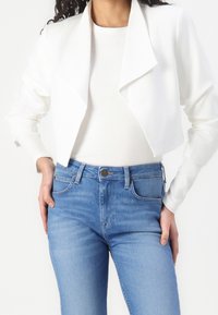 Bílá krátká bunda se širokými klopami a dlouhými rukávy, doplněná světle modrými džínami s pěti kapsami a klasickým střihem.