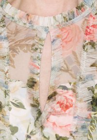 Blouse fleurie transparente avec bords à volants, dotée d'un col en forme de goutte. Couleurs comprenant rose, vert et bleu clair sur un tissu doux et translucide.