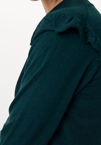 Kvinna som bär en mörkgrön stickad tröja med volangdetaljer på axeln mot en vit bakgrund, beskuren från hakan till mitten av överkroppen.