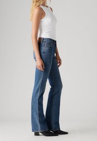 Femme portant un haut blanc sans manches, un jean bleu évasé taille haute et des bottines noires à bout pointu, debout de profil sur un fond gris.