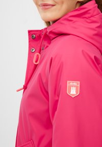 Ljus rosa vattentät jacka med huva, med en framsida med dragkedja, justerbara snoddar och en logopatch på vänster ärm.
