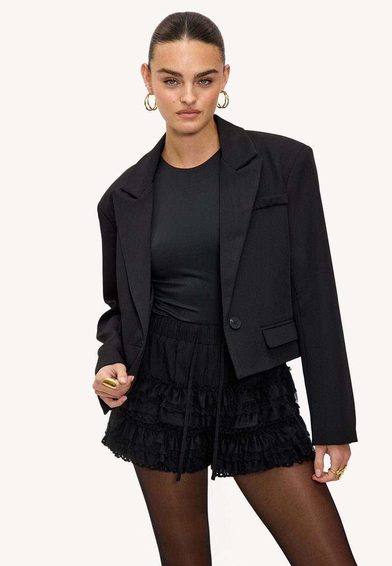 Vrouw met teruggekamd haar, draag gouden oorringen, een zwarte cropped blazer, gerimpelde zwarte shorts, doorschijnende zwarte panty’s en gouden ringen.