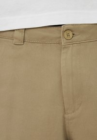 Pantalon en sergé de coton beige avec fermeture à boutons à l'avant, design à devant plat, et poches latérales ; présente une texture diagonale subtile.