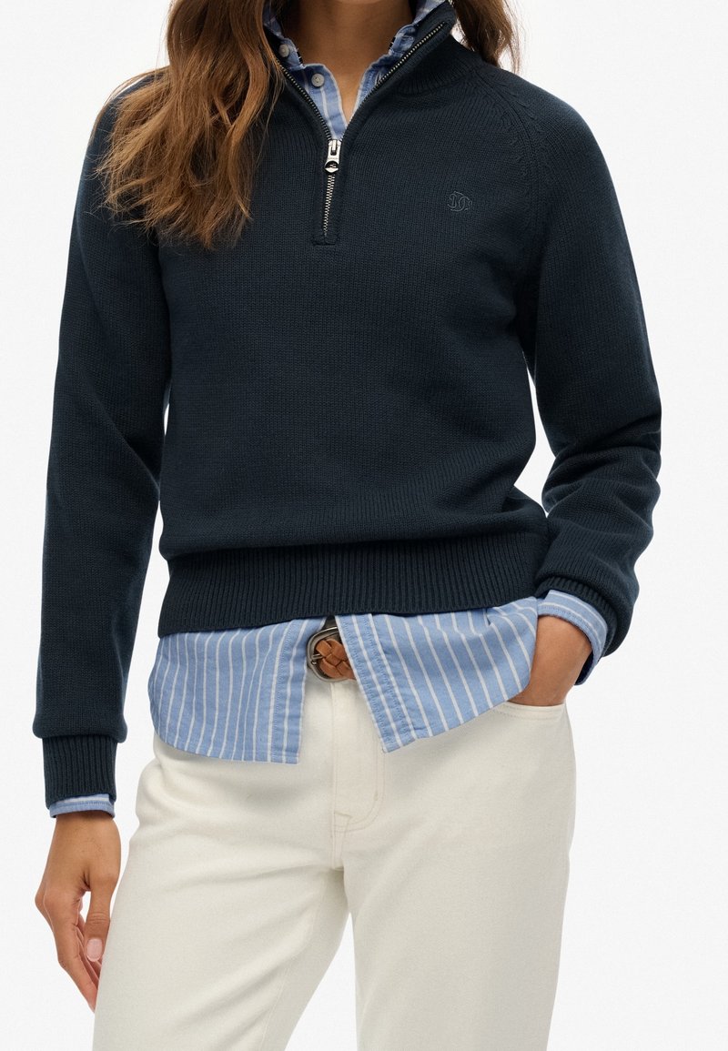 Femme portant un pull marine à demi-fermeture éclair par-dessus une chemise bleue rayée boutonnée, rentrée dans un pantalon blanc avec une ceinture tressée marron, main dans la poche.