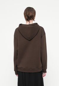 Hnedá mikina s veľkou kapucňou, vo voľnom strihu a rebrovaným lemom. Vyrobená z mäkkého materiálu, s minimalistickým dizajnom a viditeľnými švovými detailmi.