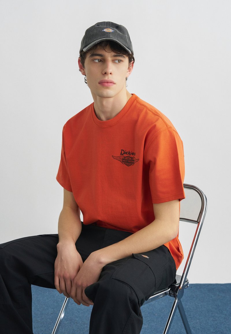 Junger Mann sitzt auf einem Metallstuhl, trägt ein oranges Dickies-T-Shirt, schwarze Cargo-Hosen und eine dunkelgraue Mütze, blickt zur Seite.