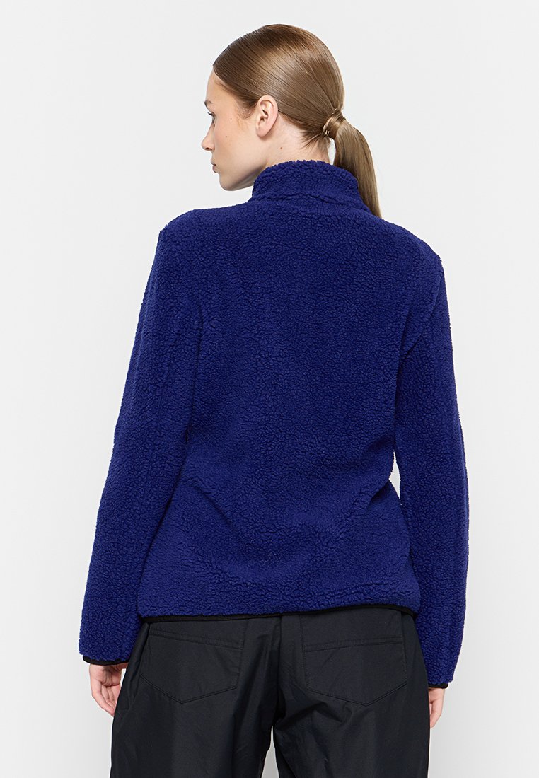 Giacca in pile blu navy con colletto alto, materiale testurizzato e design aderente, vista dal retro. Pantaloni neri visibili sotto.