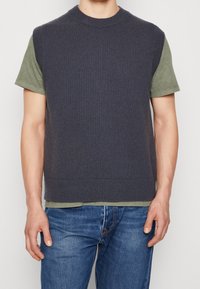 Laivastonsininen neulottu hihaton villapaita vaaleanoliivinvihreän T-paidan päällä; pyöreä pääntie, teksturoitu kangas, rento istuvuus. Yhdistetty sinisiin farkkuihin.