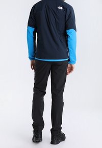 Blå och svart utomhusjacka med långa ärmar, rygglogo, kombinerad med svarta byxor och mörka skor. Slät tyg och strömlinjeformad passform.