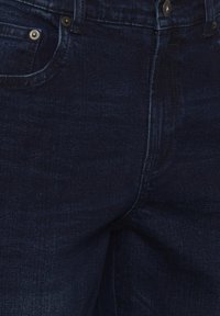Jeans de mezclilla azul oscuro con un diseño clásico de cinco bolsillos, con una sutil textura de desvanecimiento y acentos de hardware metálico.