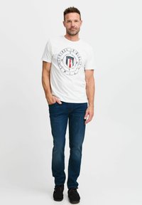 Camiseta blanca de manga corta con un gráfico circular y un diseño de escudo en rojo, azul y negro. Combinada con unos vaqueros azul oscuro y zapatos negros.