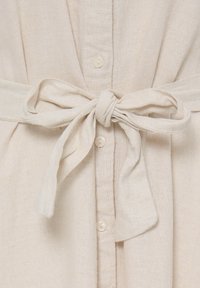 Nœud noué avec une large ceinture en tissu sur un vêtement en lin beige à boutons, montrant la texture et les détails des coutures du tissu naturel.