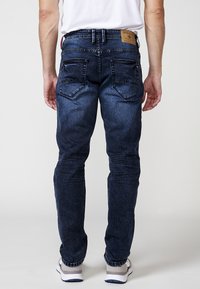 Jeans de denim albastru închis, cu o croială slim, având o etichetă din piele la talie, două buzunare pe spate și o decolorare subtilă.