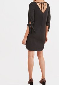 Femme portant une robe noire longueur genoux avec des manches trois-quarts nouées aux poignets et un dos ouvert avec un nœud au cou.