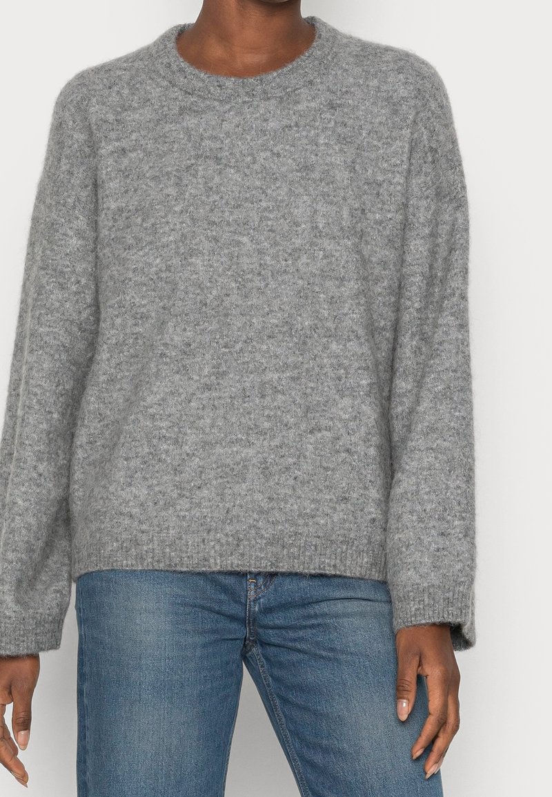 Personne portant un pull en maille gris et pelucheux à manches longues et un jean bleu sur un fond clair uni.