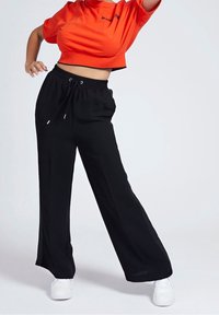 Camiseta naranja cortada con texto negro, combinada con pantalones anchos negros que cuentan con una cintura elástica y cordón. Incluye zapatillas blancas.