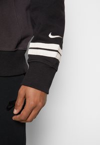Detailní záběr na ruku v černých kostkách Nike a černém rukávu s bílými pruhy a bílým logem Nike swoosh.