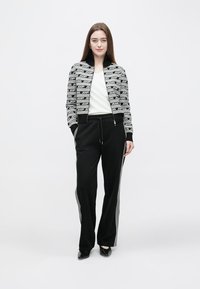 Black and white zip-up jacket with logo pattern, paired with black pants featuring grey side stripes. Both garments have a relaxed fit.