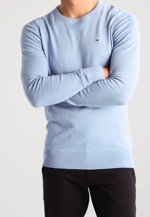 Personne portant un pull à manches longues bleu clair avec un petit logo sur la poitrine et un pantalon noir, les bras croisés au niveau du torse.