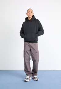 Felpa nera con cappuccio, pantaloni cargo grigi e sneakers bianche con accenti neri. Il modello si trova su uno sfondo neutro con pavimento blu.
