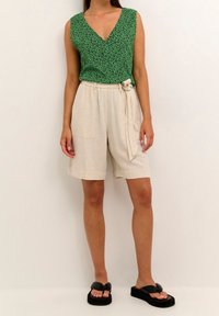 Mujer con una blusa sin mangas de flores verdes, pantalones cortos de talle alto beige con cinturón y bolsillos frontales, y sandalias de plataforma negras.