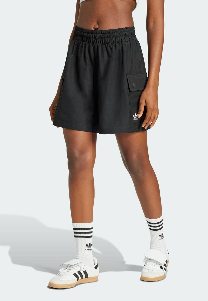 Pantalones cortos negros con cintura elástica, bolsillos laterales y un bolsillo cargo delantero. Presentan un logo blanco en la pierna. Combinados con calcetas a rayas y zapatos.