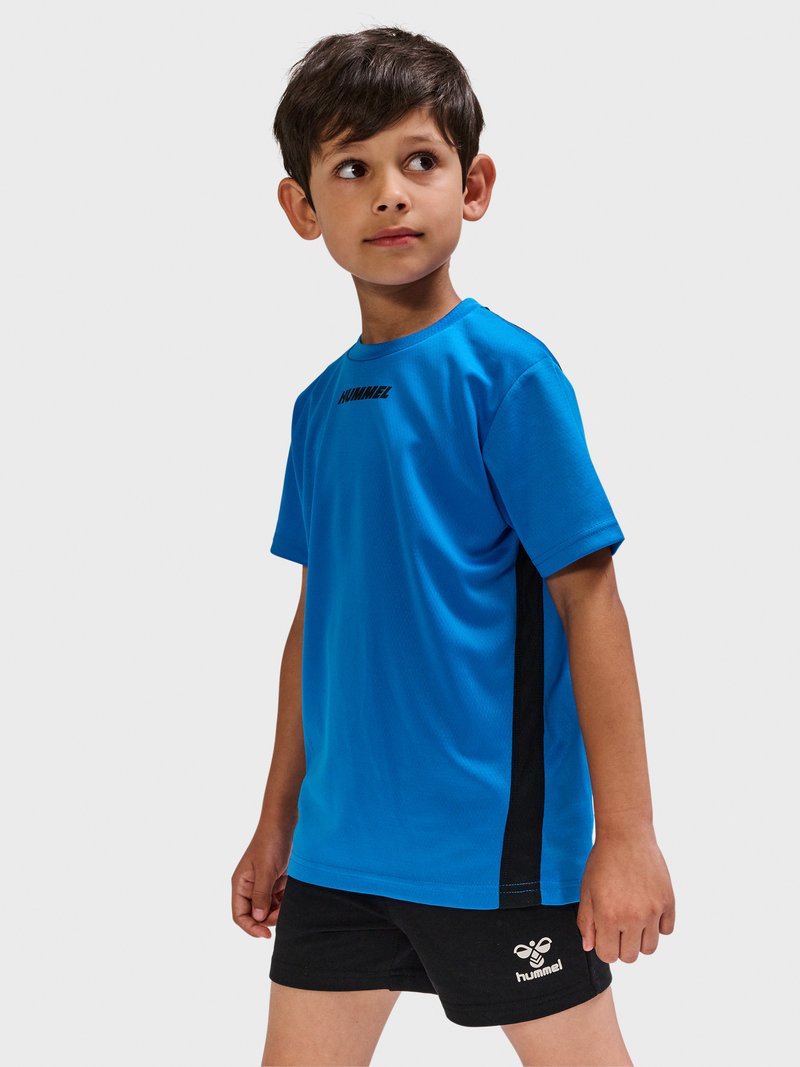 Junge steht und schaut zur Seite, trägt ein blaues Sportshirt und schwarze Shorts mit "hummel"-Logos auf einem hellgrauen Hintergrund.