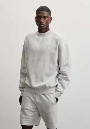 Ung mand med kort hår iført lysegrå sweatshirt og matchende shorts, stående mod en ensfarvet lys baggrund med hænderne i lommerne.