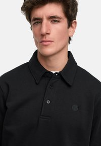 Schwarzes Poloshirt aus einem Baumwollmischgewebe, mit Kragen, einer Drei-Knopfleiste und einem kleinen gestickten Logo auf der Brust.